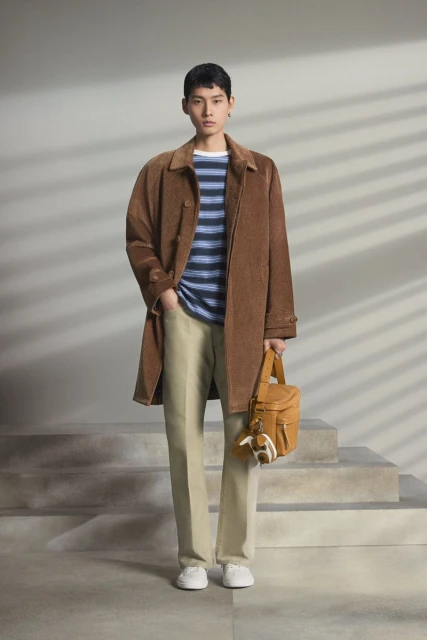 Male fashion model poses on stairs indoors wearing a brown coat, striped shirt, khaki pants, white shoes, and holding a purse.