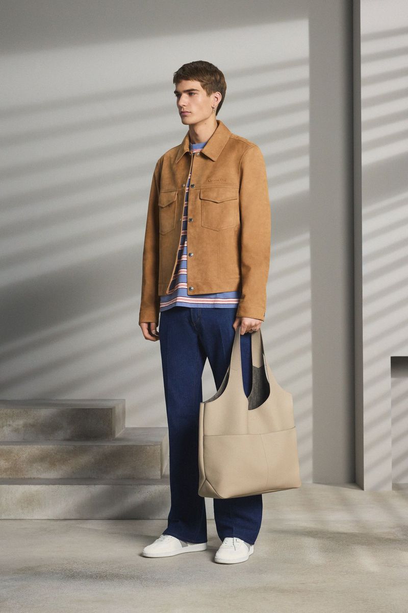 Model wearing tan suede jacket and blue trousers holding a beige tote bag against a structured wall with window light shadows