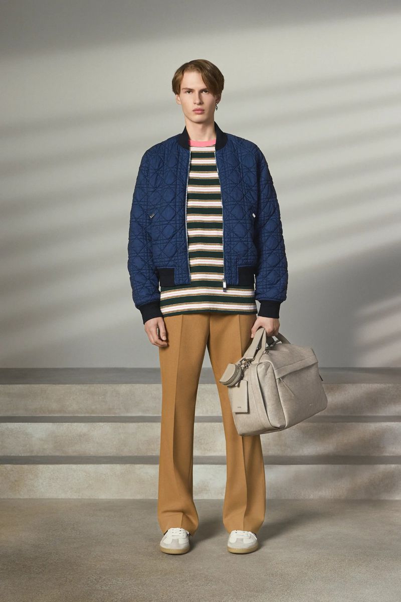 Man poses in a blue quilted jacket, striped shirt, and tan pants, holding a bag, on a neutral background with stairs, in a studio.