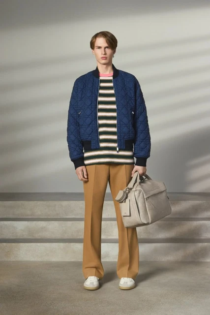 Man poses in a blue quilted jacket, striped shirt, and tan pants, holding a bag, on a neutral background with stairs, in a studio.