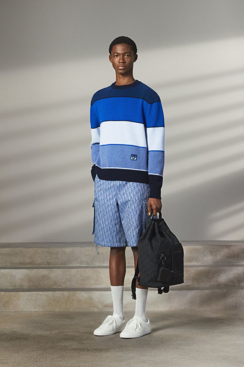 Model standing on steps wearing a blue striped sweater, patterned shorts, and holding a dark backpack.