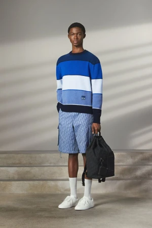 Model standing on steps wearing a blue striped sweater, patterned shorts, and holding a dark backpack.