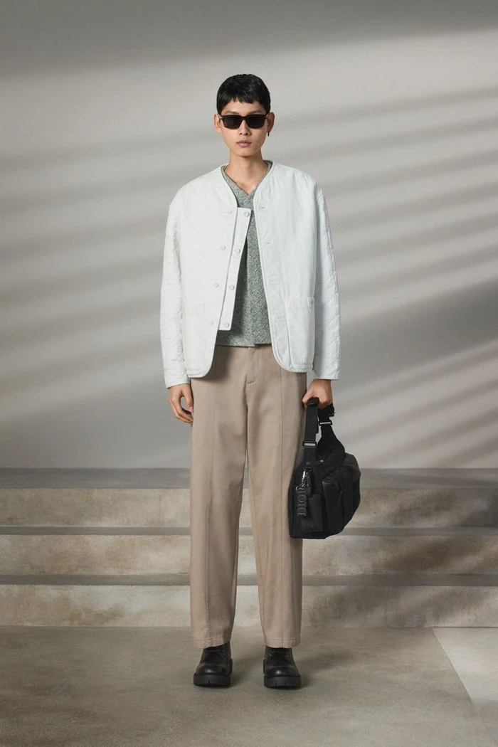 Model wearing a light grey quilted jacket, brown trousers, and sunglasses while holding a black shoulder bag.