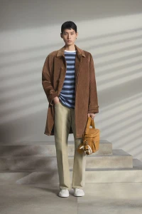 Model standing in a brown coat holding a bag against a grey seamless studio backdrop with slatted light patterns