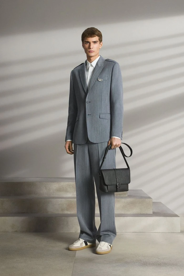 Model wearing a grey suit and holding a black messenger bag standing before light-streaked architectural steps.