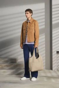 Model wearing tan suede jacket and blue trousers holding a beige tote bag against a structured wall with window light shadows