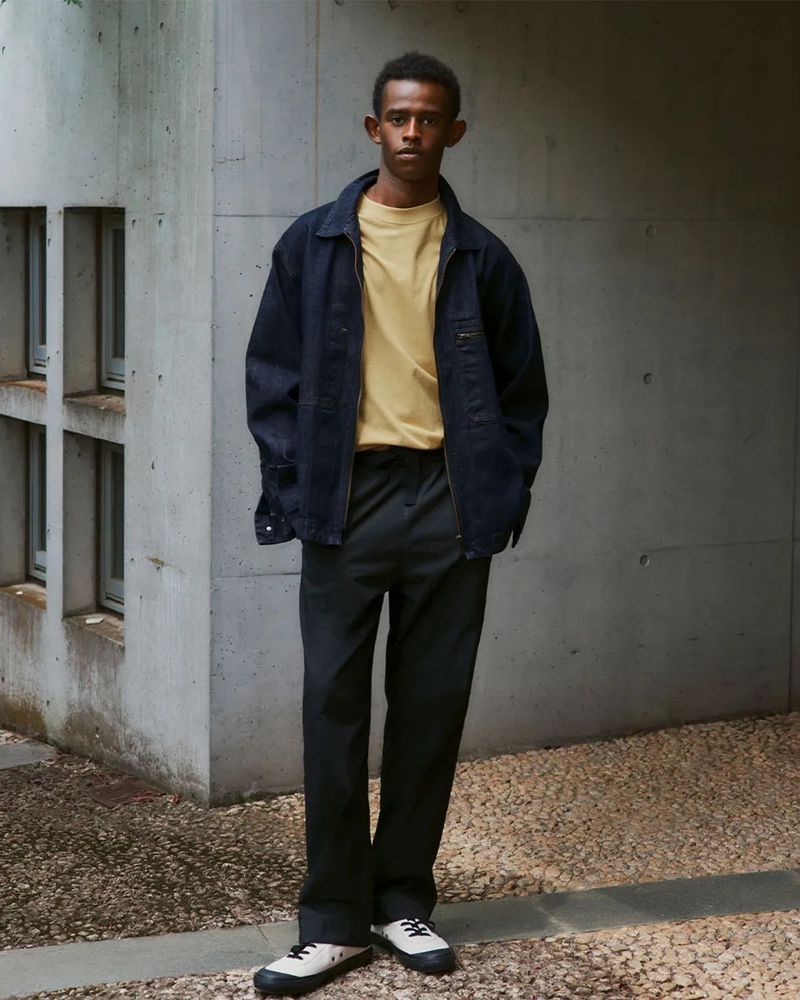 Young man in denim jacket and black pants stands against concrete building. Earthy neutral tones, natural light, minimalist fashion.