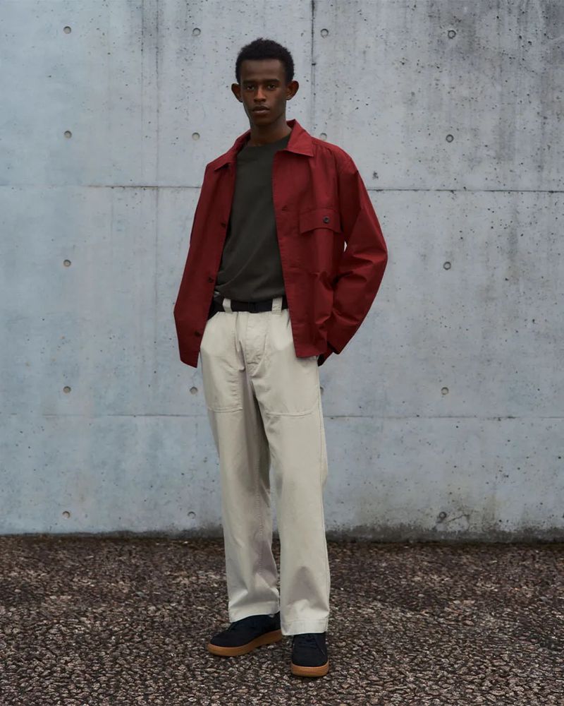 Full shot of a young adult man in casual fashion: red work jacket, green tee, beige pants, and black sneakers posing against a concrete wall.