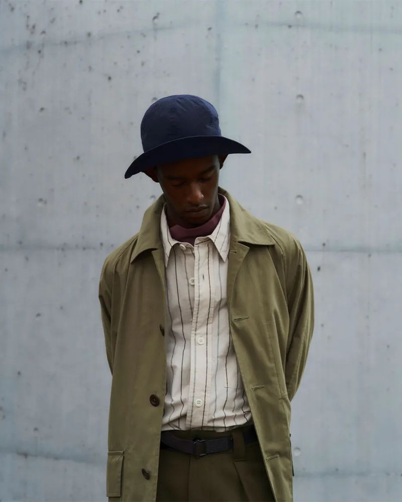 Studio shot of a stylish Black male model wearing a blue hat, striped shirt, and trench coat, posing against a gray concrete wall.