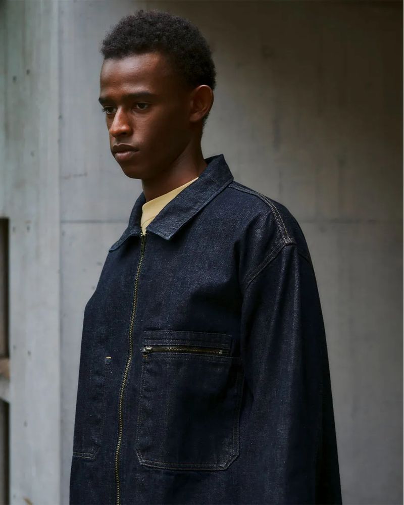 Model wearing a dark denim jacket with contrast stitching against a concrete architectural background.