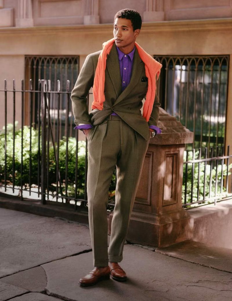 Man in olive suit stands in front of a building wearing a purple shirt and orange sweater around his neck in an urban setting.