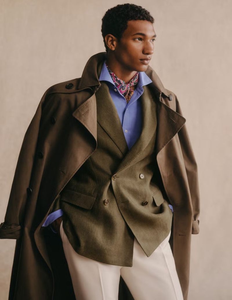 Man in a green suit and trench coat looking to the side against a neutral background in a studio shot. Fashion editorial.