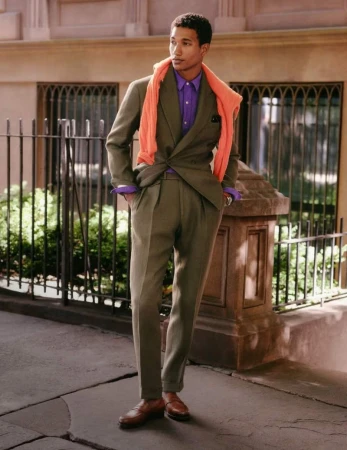 Model wearing an olive suit, purple shirt, and orange sweater draped over shoulders outdoors on a city sidewalk