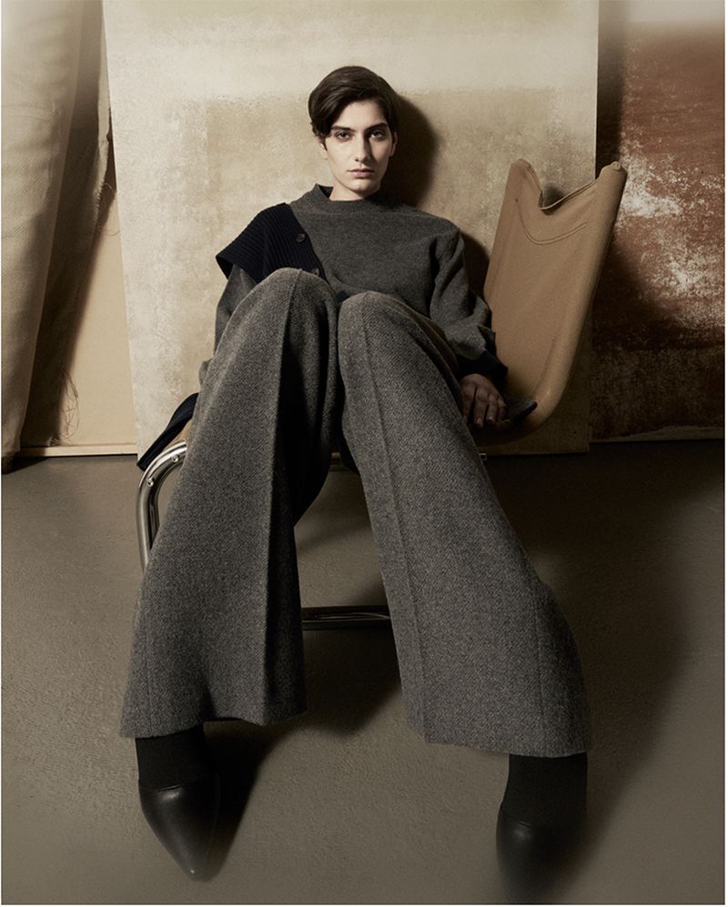 Fashion editorial shot of a woman in a gray wool outfit sitting in a tan chair against a neutral backdrop in a photo studio.