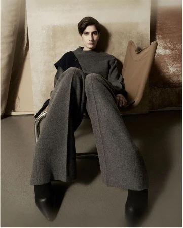 Fashion editorial shot of a woman in a gray wool outfit sitting in a tan chair against a neutral backdrop in a photo studio.
