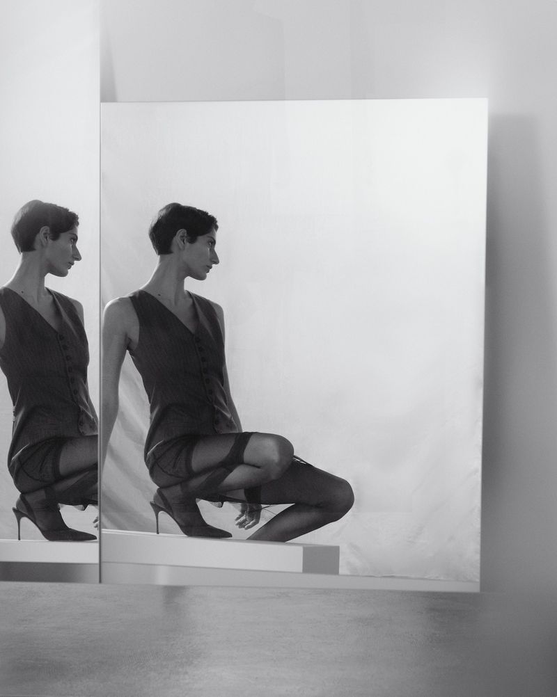 Monochrome portrait of a model crouched on a block, reflected in a large mirror for Eudon Choi Prefall 2025.