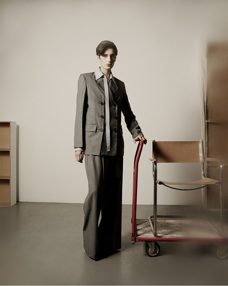 Fashion photo of a woman in a grey suit standing next to a red hand truck in a neutral indoor setting. Editorial style portrait.
