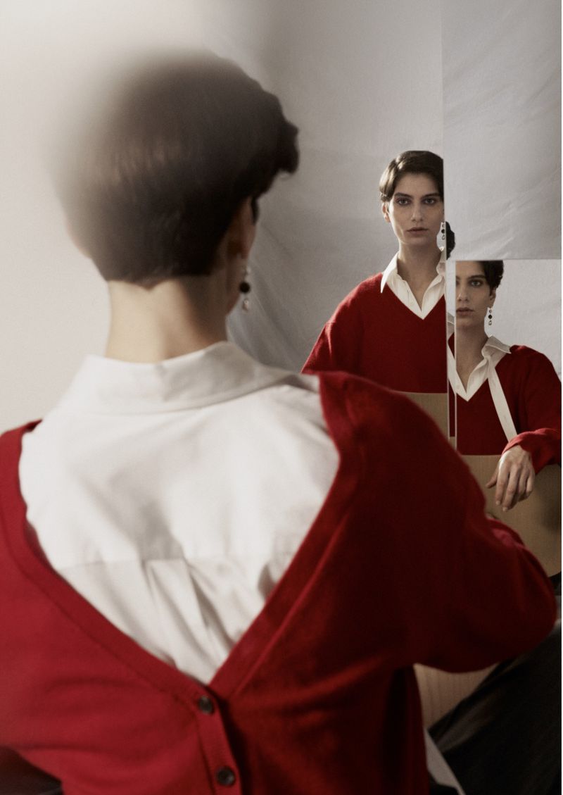 A woman in a red cardigan and white blouse poses in front of a shattered mirror, her reflection visible in the fragmented pieces.