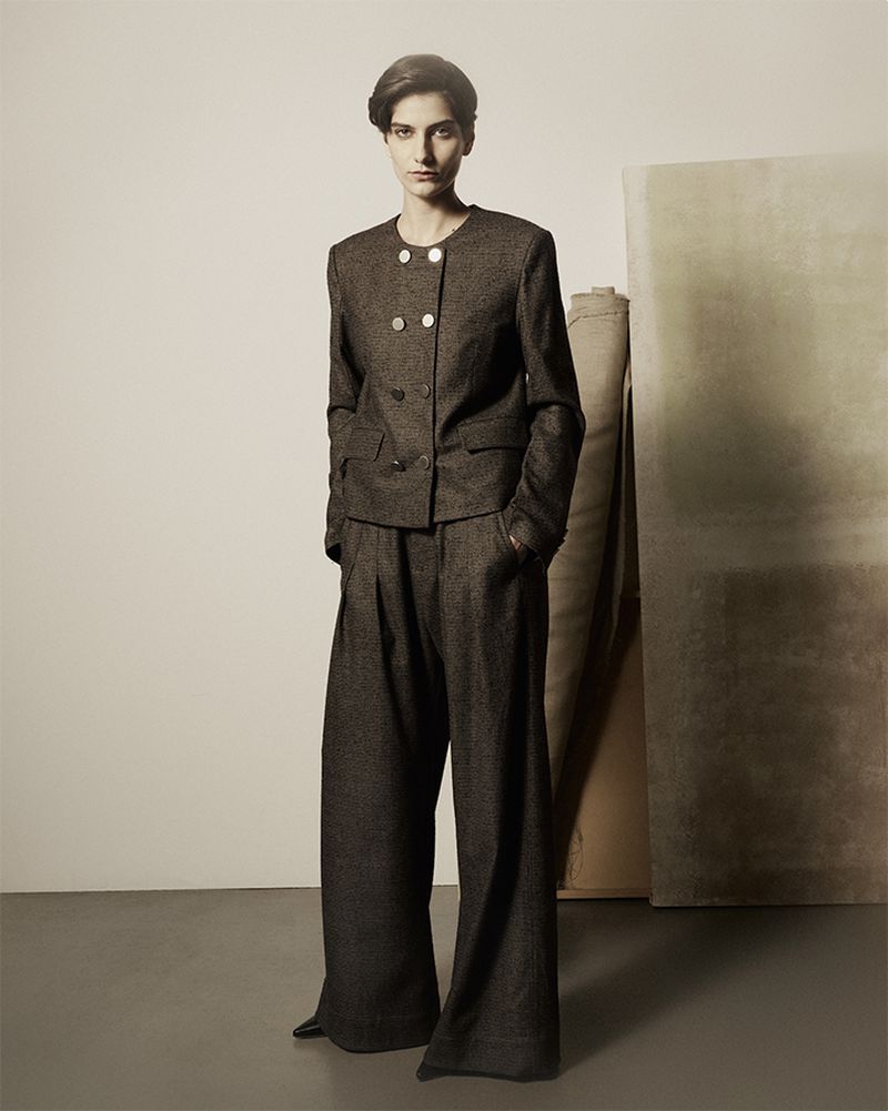 A woman poses in a tailored, dark tweed suit with a double-breasted jacket and wide-legged trousers, against a neutral backdrop.