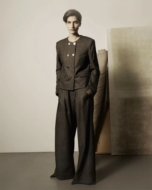A woman poses in a tailored, dark tweed suit with a double-breasted jacket and wide-legged trousers, against a neutral backdrop.