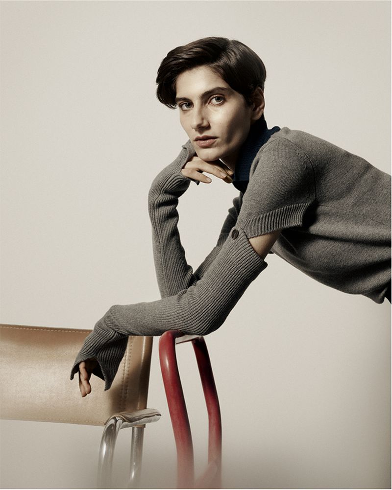 Portrait of a woman with short hair wearing a grey sweater, leaning on the back of two chairs in a minimalist photo studio setting.