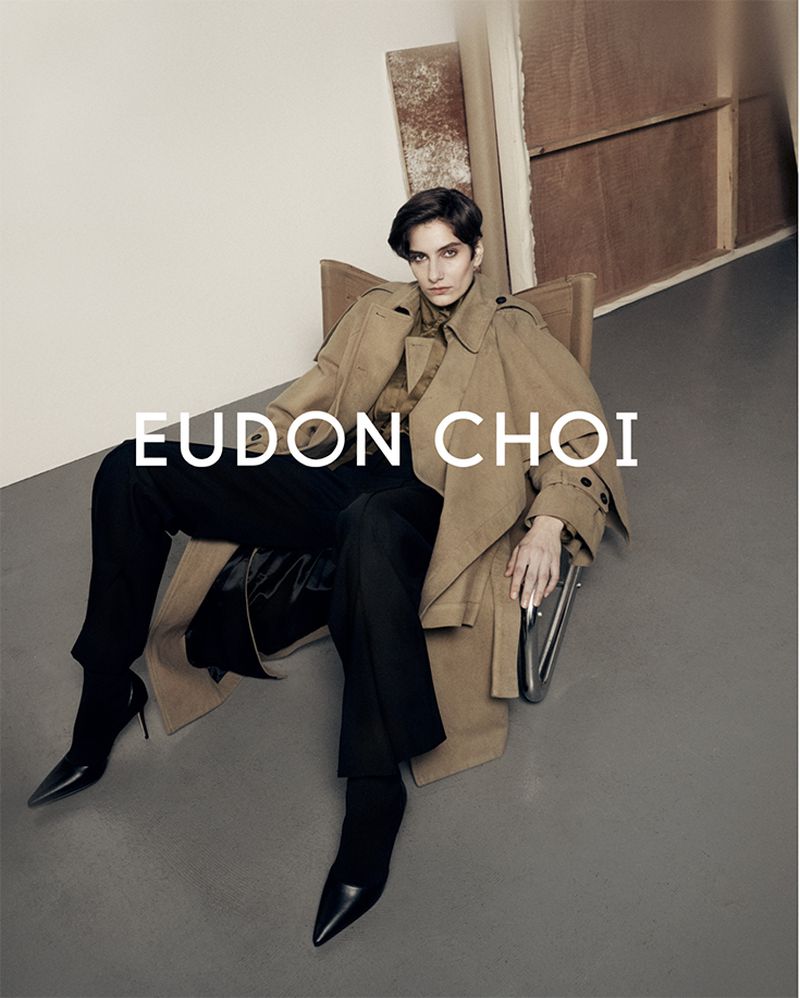 A fashionable woman with short dark hair, wearing a tan trench coat and black pants, sits in a chair in a minimalist photo studio.