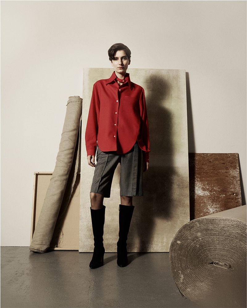 A woman in a red blouse and grey shorts stands indoors surrounded by beige and brown props in a fashion photography studio setup.