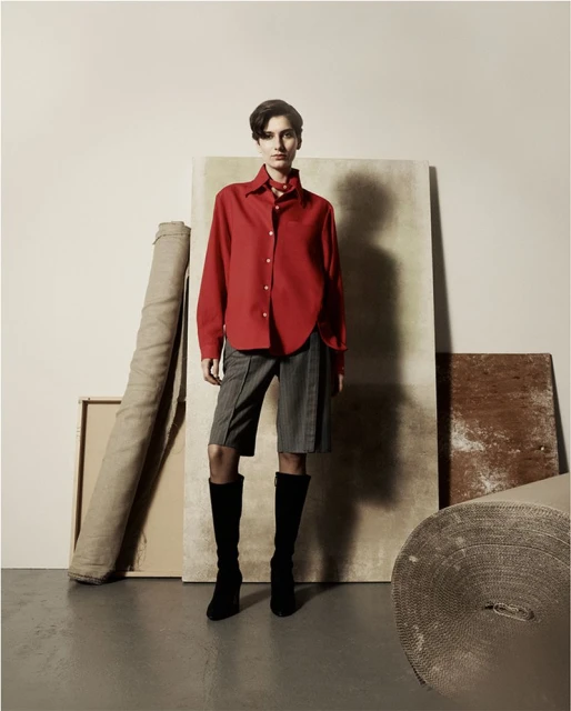 A woman in a red blouse and grey shorts stands indoors surrounded by beige and brown props in a fashion photography studio setup.