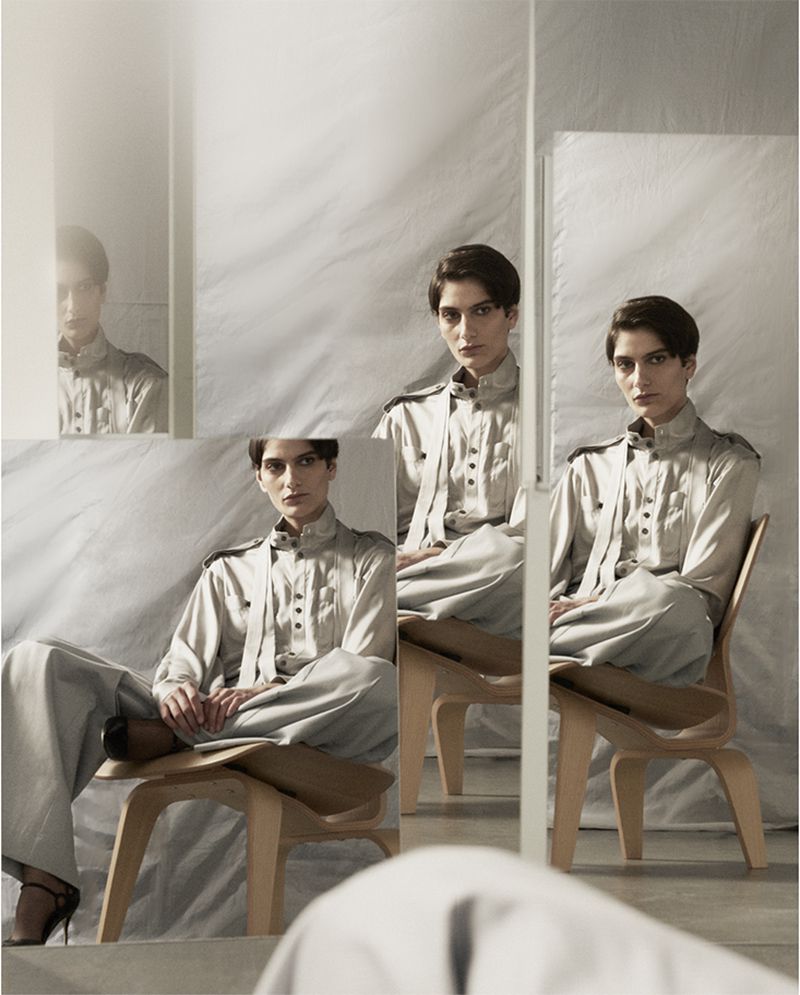 Elegant woman sits in a wood chair in front of mirrors, wearing a grey collared shirt and pants, with a neutral expression in a studio.