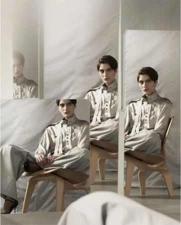Elegant woman sits in a wood chair in front of mirrors, wearing a grey collared shirt and pants, with a neutral expression in a studio.