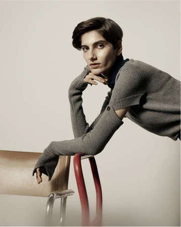 Model leaning on a red and chrome chair wearing a grey knit top with cutout sleeves for Eudon Choi
