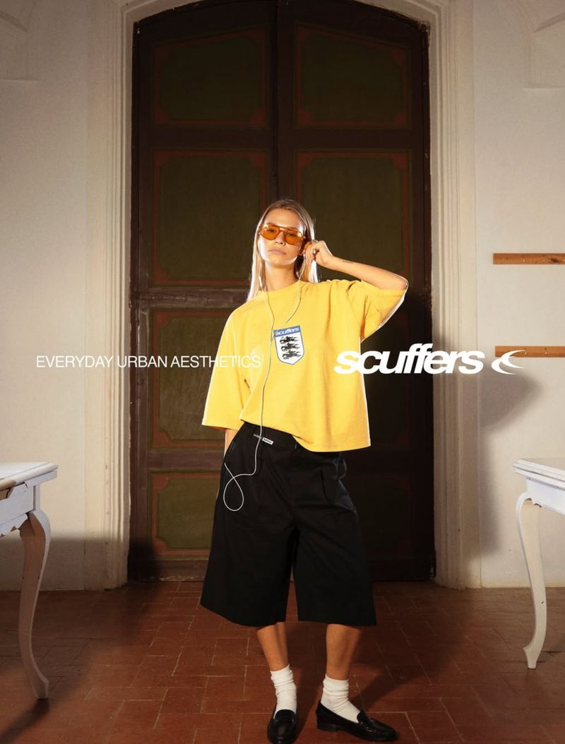 Woman in yellow tee and black shorts poses in doorway with headphones and sunglasses. Photo features white desks and brown floor.