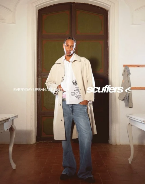 A tall, slender male fashion model in a trench coat, jeans, and t-shirt poses in front of a vintage door in an indoor setting.