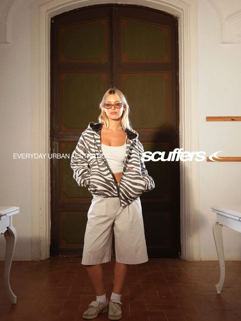 A young woman poses in front of a door, wearing a zebra print jacket, white top, beige shorts, white socks, and white velcro shoes.