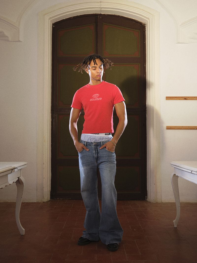 Young adult male in red shirt and jeans standing in a room with dark wood door and tile floor, flanked by two white tables.