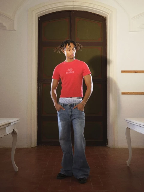 Young adult male in red shirt and jeans standing in a room with dark wood door and tile floor, flanked by two white tables.