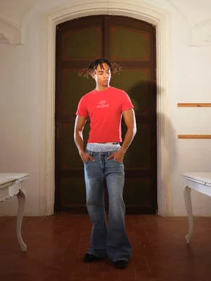 Young man standing indoors wearing a red t-shirt and blue jeans with hands in pockets