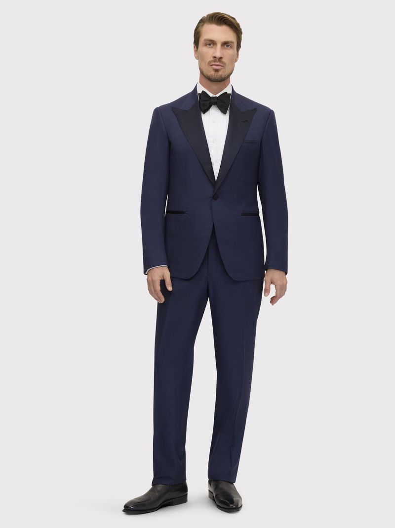 Full shot of a man in a formal navy blue suit with a black bow tie and black dress shoes, standing against a neutral grey background.