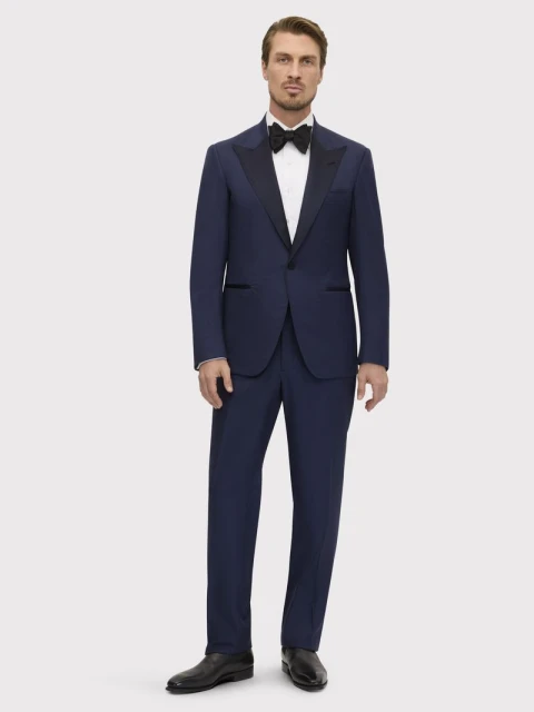 Full shot of a man in a formal navy blue suit with a black bow tie and black dress shoes, standing against a neutral grey background.