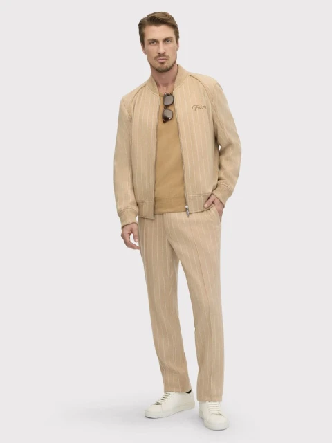Full-length portrait of a man in a tan striped suit, tan shirt, and white sneakers posing against a neutral background in a studio setting.