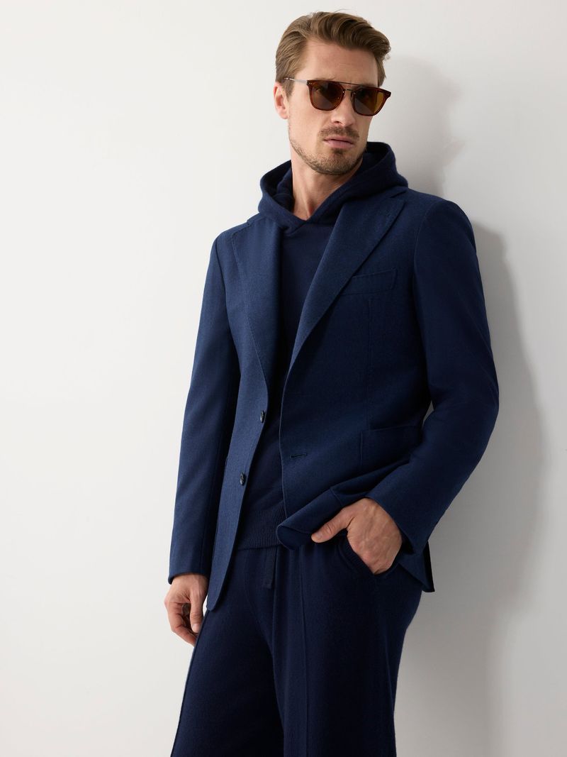 Man wearing a navy blazer and hoodie with sunglasses standing against a white seamless background