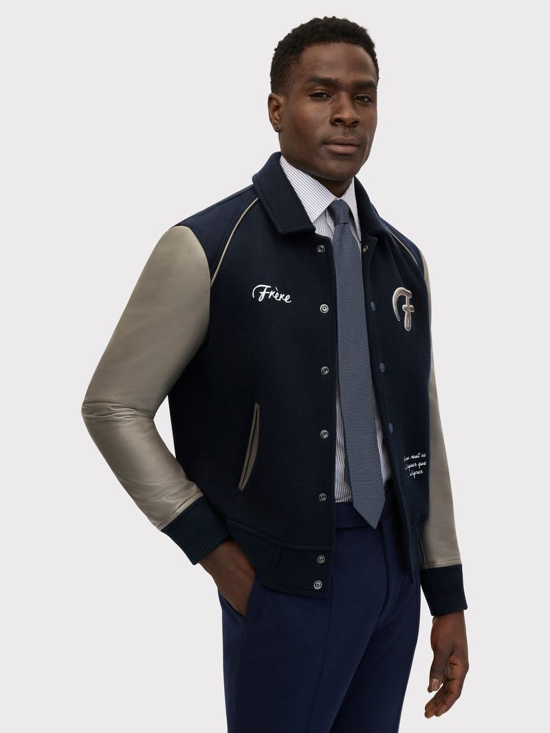 A dark-skinned man wearing a navy and gray letterman-style jacket, shirt, and tie, standing in front of a light gray background.