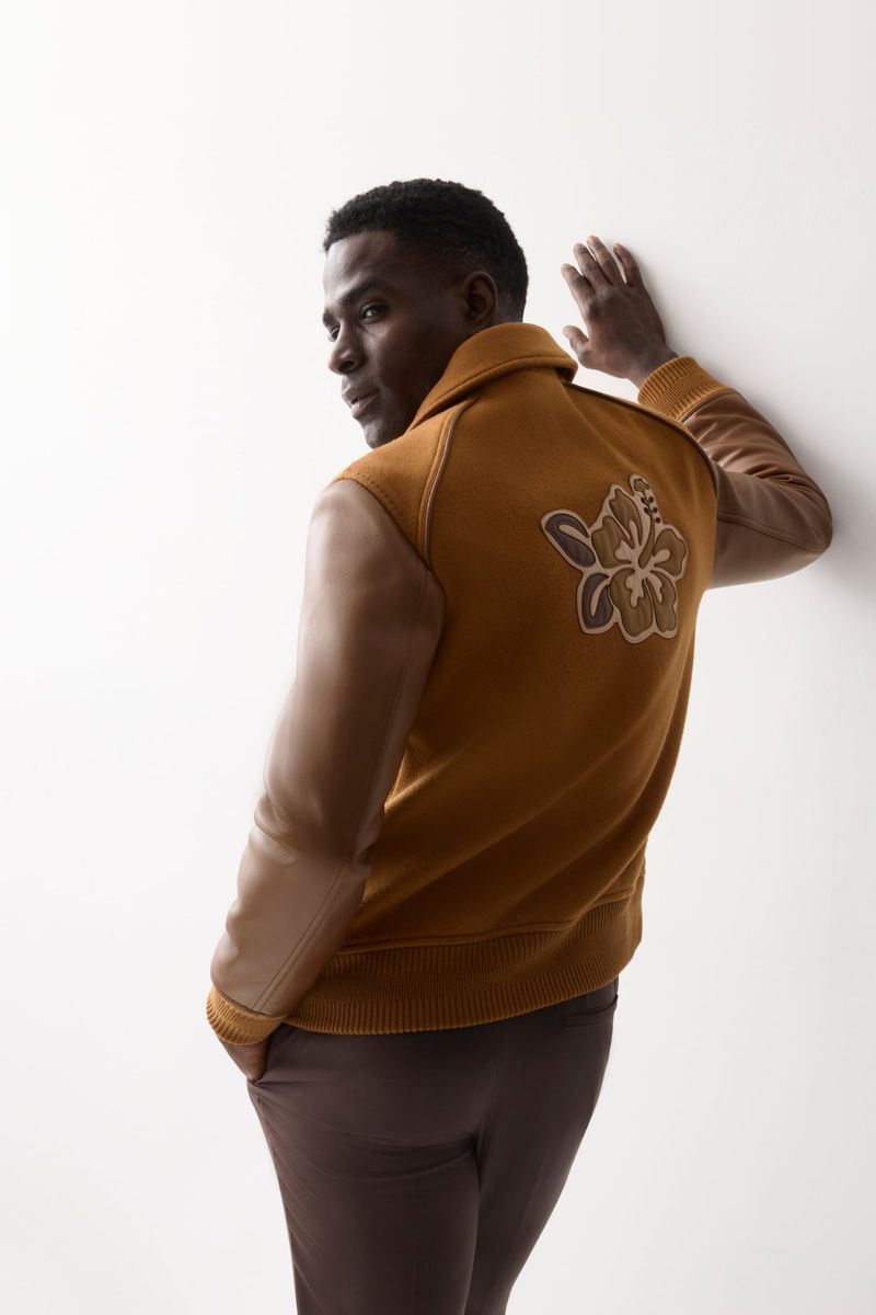 Adult male wearing an amber-colored jacket with a floral design stands against a white wall in a fashion studio setting.