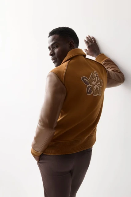 Adult male wearing an amber-colored jacket with a floral design stands against a white wall in a fashion studio setting.