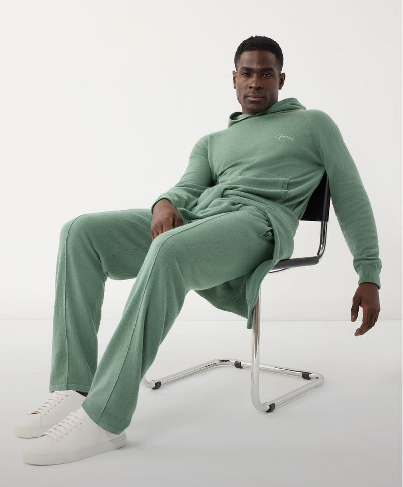 Man in a light green hooded sweatsuit sitting on a metal chair in a brightly lit studio. He's wearing white sneakers.