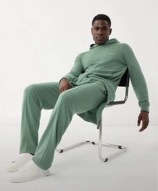 Man in a light green hooded sweatsuit sitting on a metal chair in a brightly lit studio. He's wearing white sneakers.