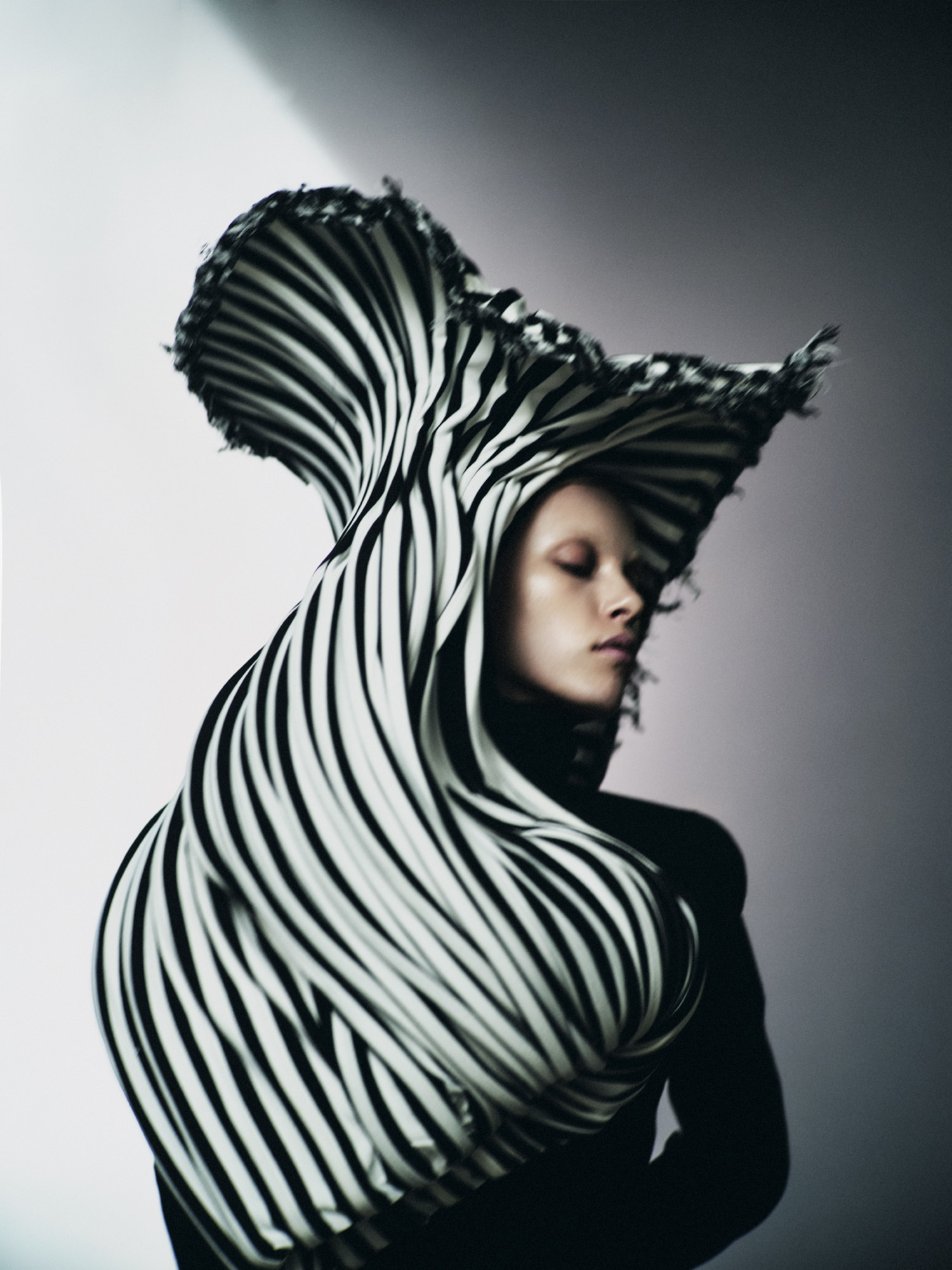 Striking fashion portrait of a woman with pale skin draped in black-and-white striped fabric, eyes closed, against a gradient grey background.