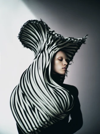 Striking fashion portrait of a woman with pale skin draped in black-and-white striped fabric, eyes closed, against a gradient grey background.