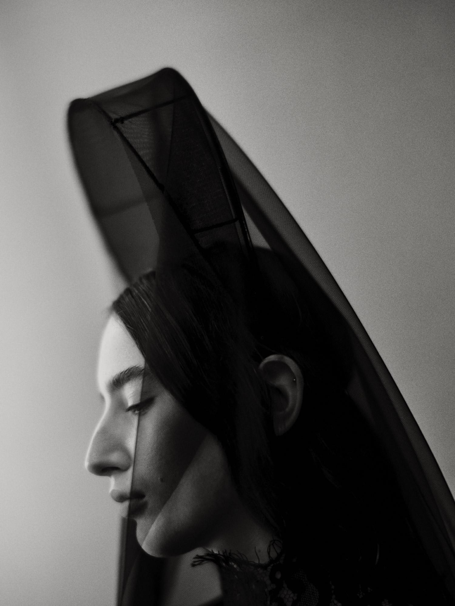 Monochrome portrait of a woman in profile wearing a black lace top and a dramatic black veil. Neutral backdrop and soft lighting.