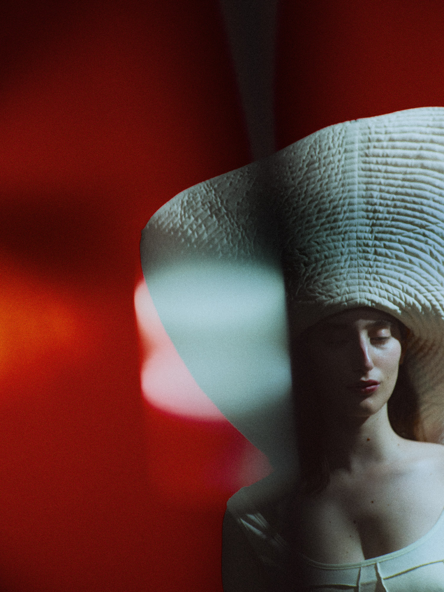 Woman in a large straw hat poses in front of a vibrant red backdrop, illuminated by soft, diffused lighting creating a surreal atmosphere.